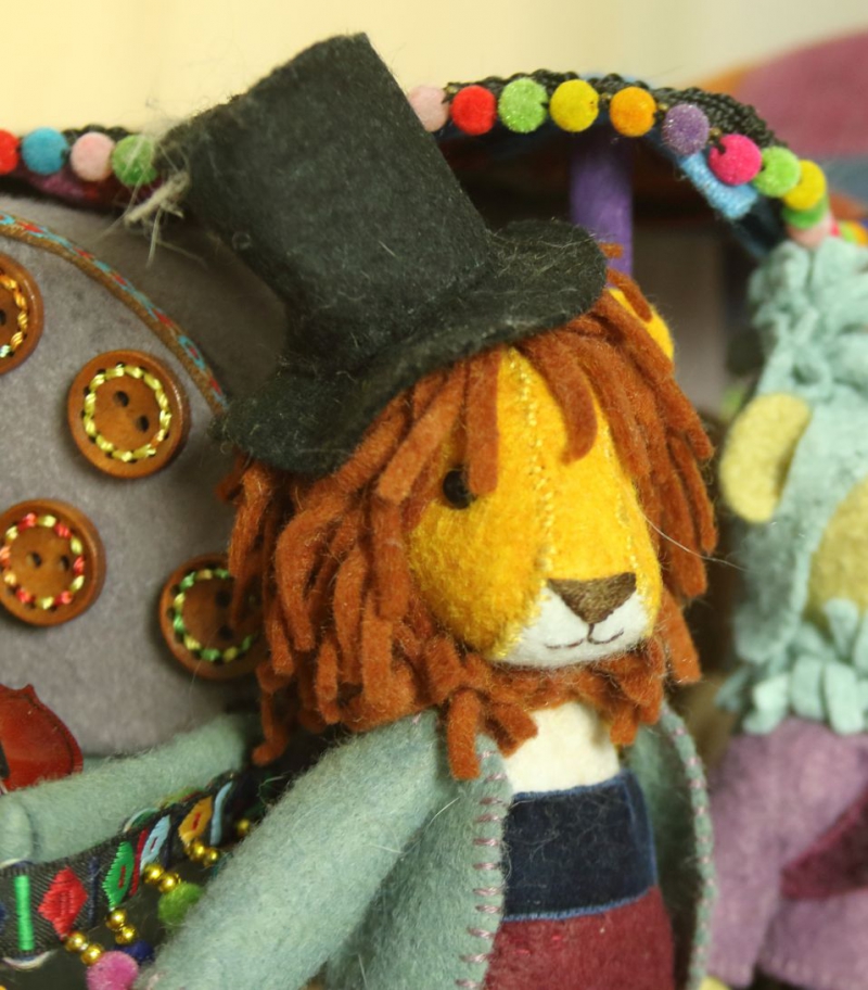An image of a doll in Barbara Schoenoffâ€™s assemblage, Rendezvous. This doll is Giuseppe the Lion, standing in front of his tent. As an adult male lion, he has a mane. Being a humanized animal, he also has a top hat on his head a light jacket.