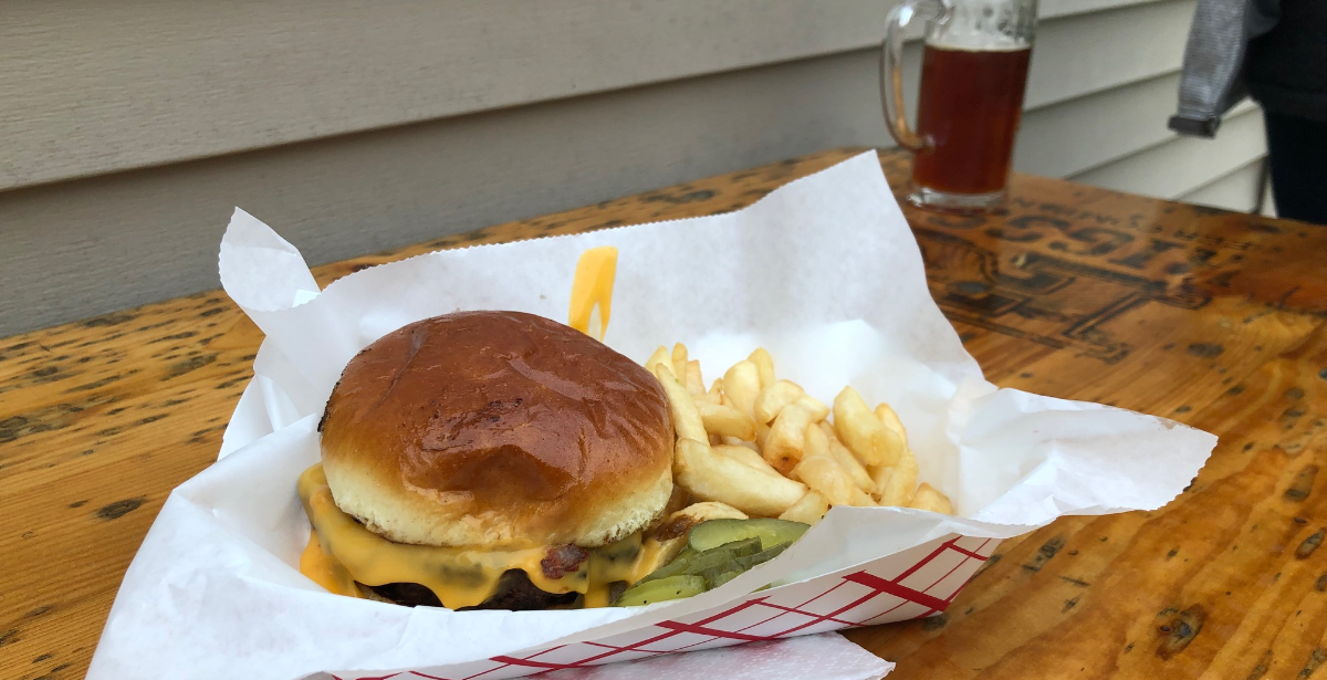 On a brown table at Riggs, there is a burger with dripping cheese beside fries in a white parchment paper lined red-and-white paper boat. In the background on the wooden Riggs table, there is a mostly full pint of beer. Photo by Alyssa Buckley.