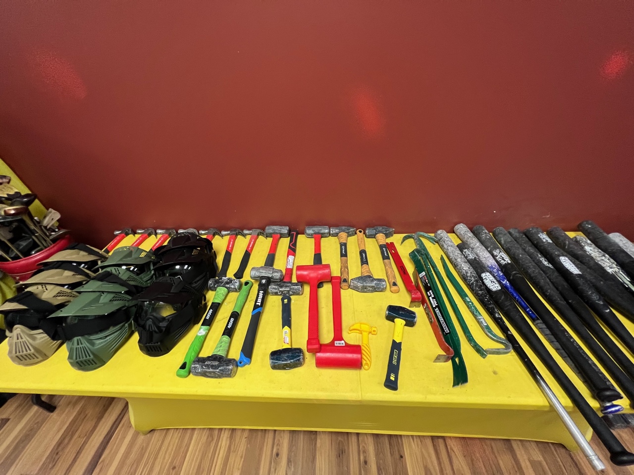 On a yellow table, there are military style helmets, sledge hammers, hammers, crowbars, and baseball bats neatly arranged. Photo by Alyssa Buckley.