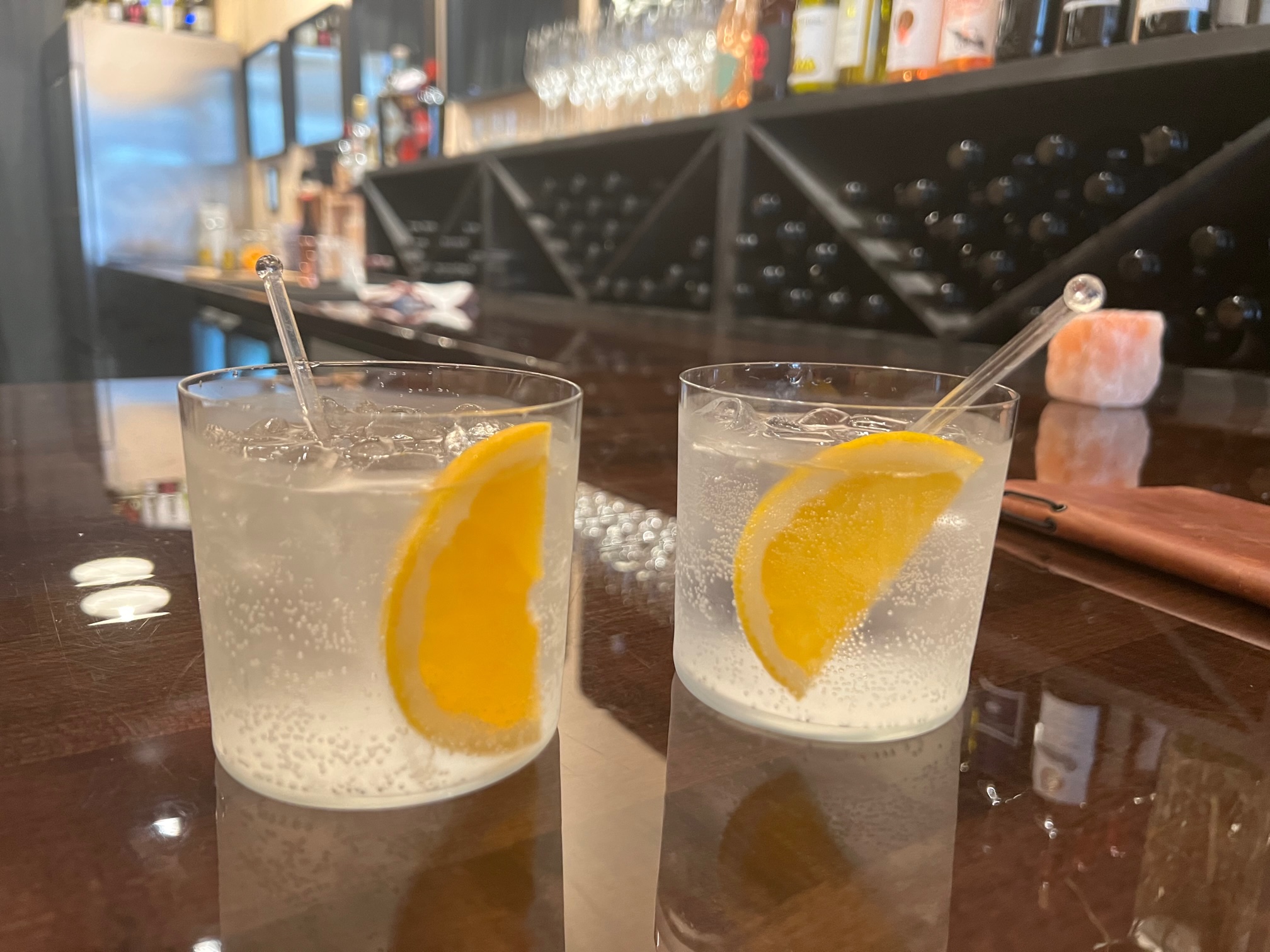 On a dark wooden bar at Ladro Enoteca, there are two gin and tonics in a clear half tumbler with a slice of orange and a clear stir stick. Photo by Alyssa Buckley.