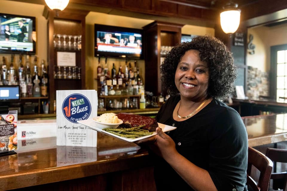 Owner of Neil St. Blues in Champaign Gayle Starks holds a platter of Southern cuisine in front of their award. She is smiling at the camera in the bar area of the Downtown Champaign restaurant. Photo from Neil St. Blues' Facebook page.