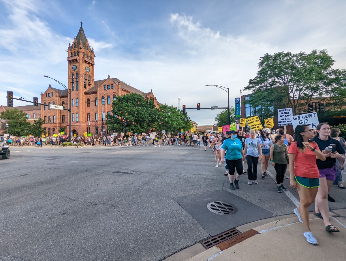 A long line of protesters holding signs extends from a reddish brick building across an intersection.
