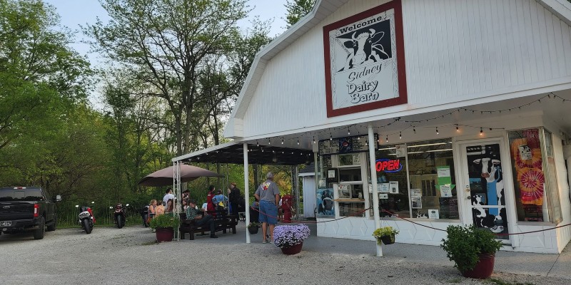 A view of the checkout window at the Sidney Dairy Barn. The barn is white, there is a gravel parking lot around it. Picnic tables are to the left of the building. Photo from Sidney Dairy Barn Facebook page.
