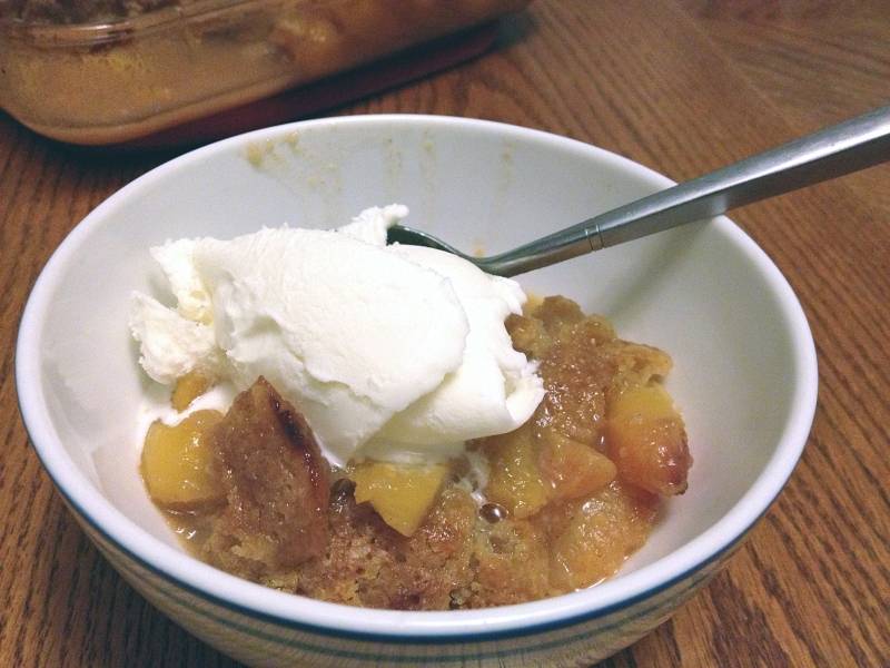 Peach cobbler with a scoop of Prairie Fruits Farm gelato served in a while bowl. A metal spoon is in the bowl. The bowl is on a brown wood table. Photo by Jessica Hammie.