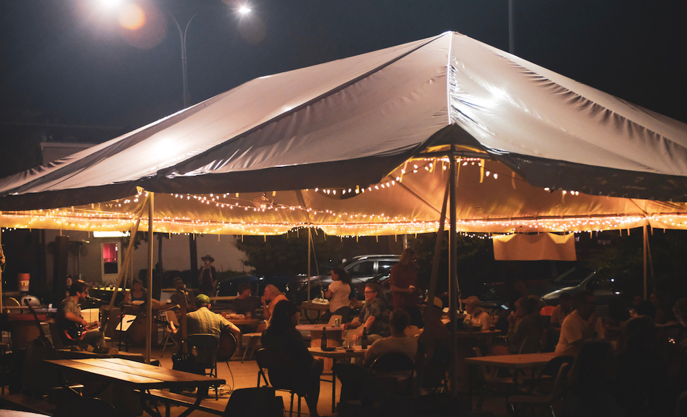 Image of the tent outside of Rose Bowl Tavern. String lights hang, patrons sit at chairs under the tent.