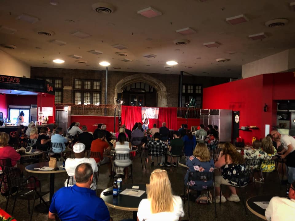 A crowd of people viewed from behind, seated, looking at a stage with red curtains. Someone is sitting on a stool on the stage, but is obscured by a bright spotlight.. There are red walls on either side of the curtains. Photo from the Facebook event page.