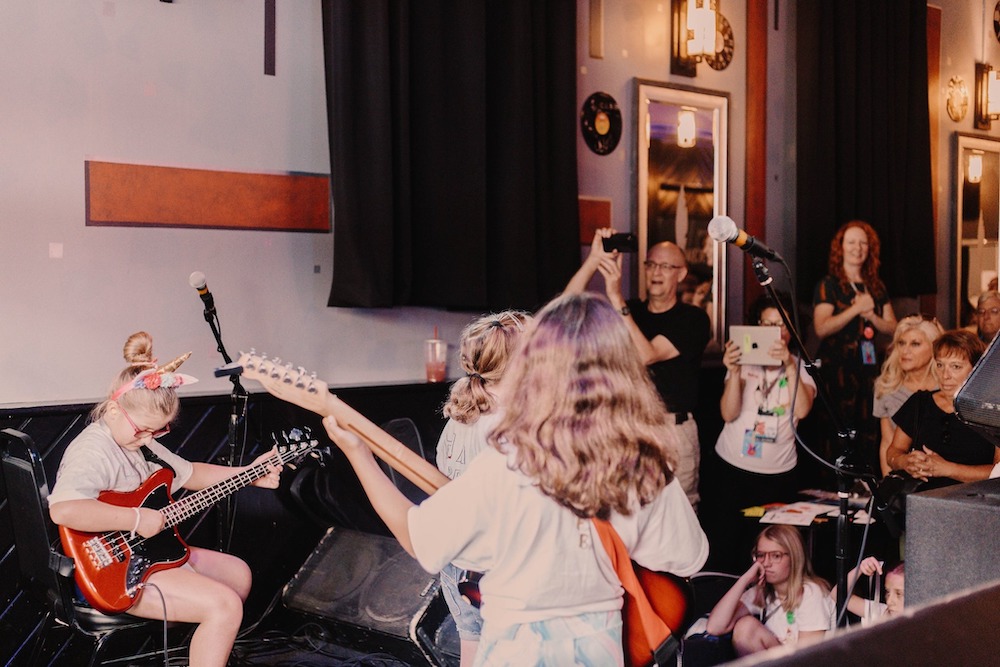 A group of young girls play instruments including bass and guitar on a stage, as an audience cheers them on.