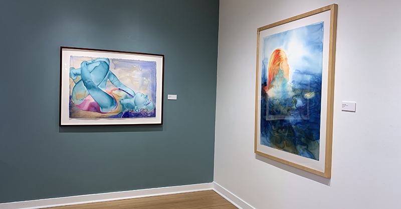 On the left, against a blue wall, a painting with a woman lying down holding her knees to her chest. On the right, a painting of a orange hued woman emerging from a deep blue body of water against a white wall. 
