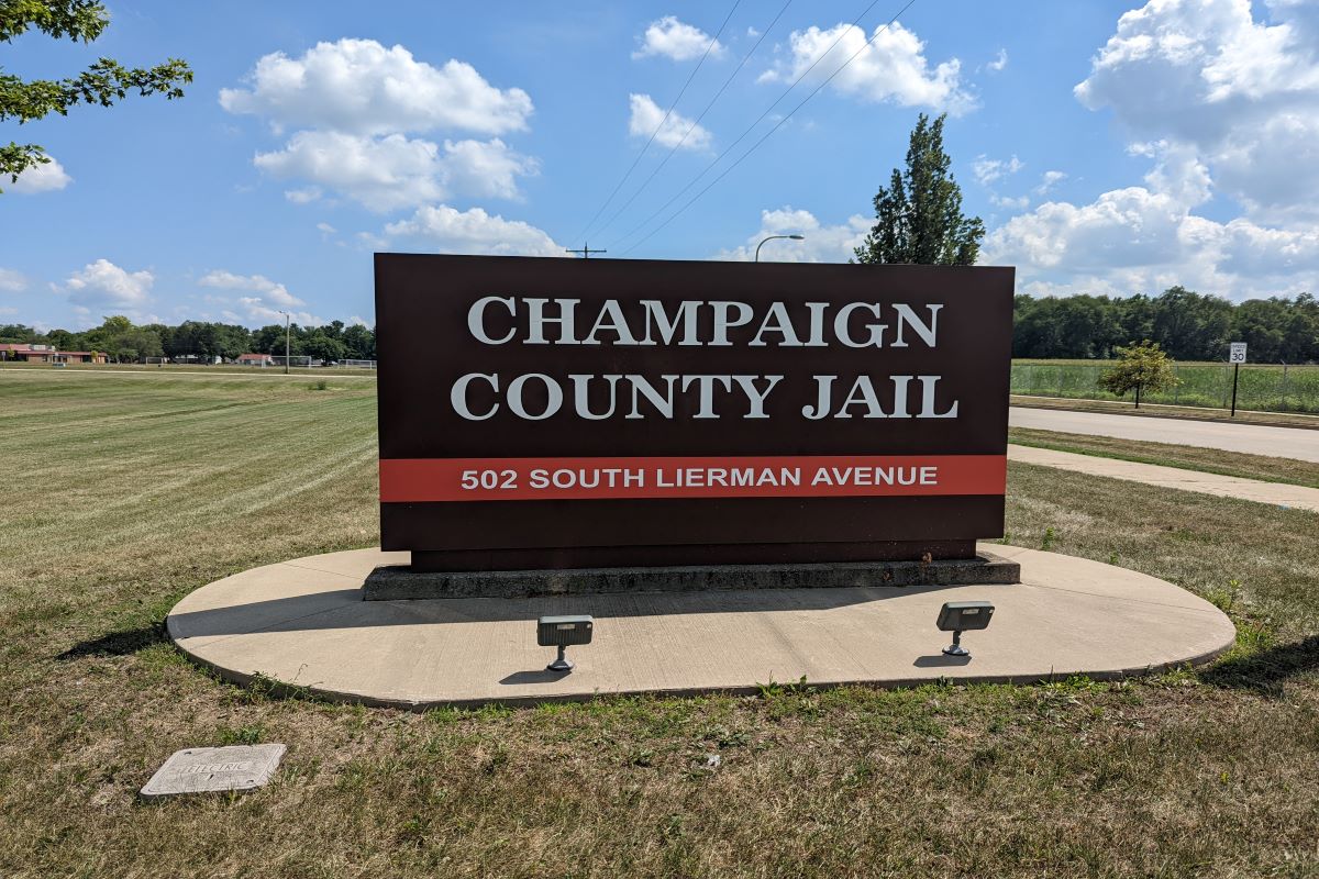 The sign for the Champaign County Jail. It is a dark brown sign with white letters and a red stripe with the address across the bottom. In the photo the sign is mounted to a concrete slab. It is a sunny day, with a blue sky and a scattering of puffy clouds.