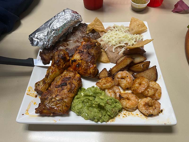 A white, rectangular plate holds a crispy, charred chicken thigh and leg, seven pieces of grilled shrimp, a slice of carne asada, a foil-wrapped cylinder of tortillas, a mound of refried beans with cheese on top and tortilla chips sticking out to the sides, and a scoop of bright green guacomole. Photo by Julia Freeman.