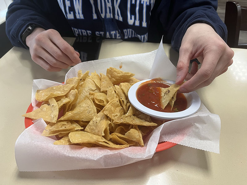 A red plastic basket lined with parchment paper and filled with tortilla chips sits on a table. A person wearing a navy-blue sweatshirt dips a chip into a bowl of red salsa nestled next to the chips. Photo by Julia Freeman.