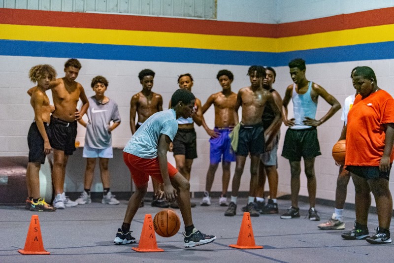 A Black teen in a blue t-shirt and red shorts dribbles a basketball between his legs, while weaving through orange cones. A line of Black teens watch. Photo by J. Sidney Malone. 
