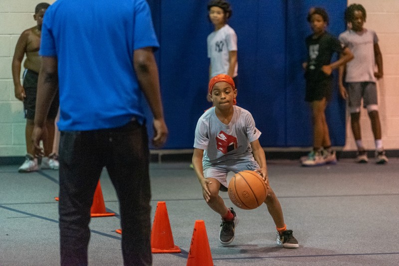 A young, Black boy in a gray shirt and an orange do-rag is crouched low, dribbling a basketball through a set of orange cones. Photo by J. Sidney Malone.