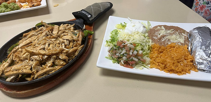 A cast iron skillet piled with thin strips of chicken rests on top of a wooden holder. To the right of the skillet, a white, rectangular plate holds mounds of lettuce, cheese, sour cream, pico de gallo, refried beans with chees on top, seasoned rice, and a foil-wrapped cylinder of tortillas. Photo by Julia Freeman.
