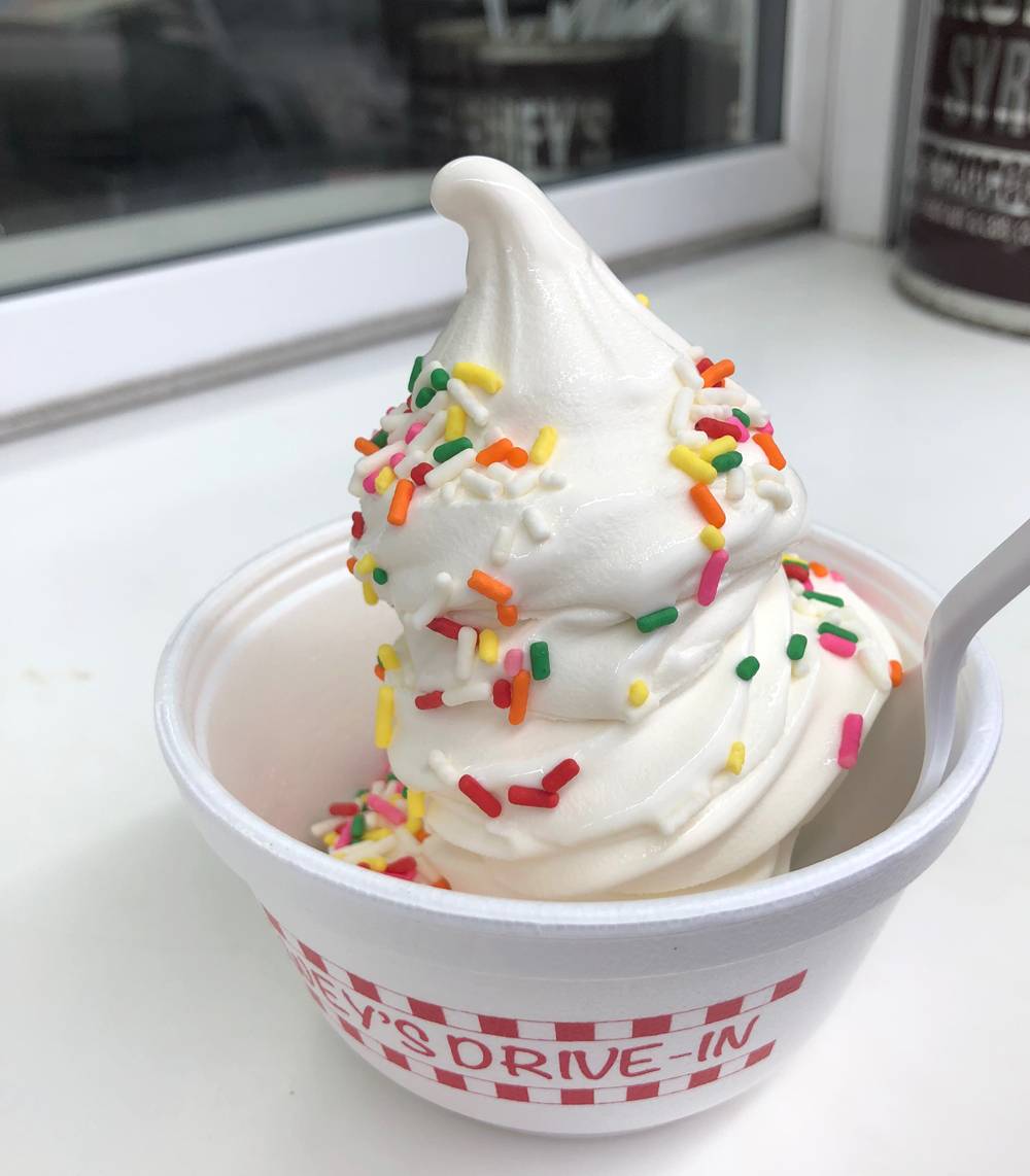Soft serve vanilla ice cream with rainbow sprinkles is in a white styrofoam cup. The cup has red lettering that says 