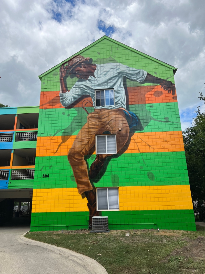 A brightly colored mural covers the entirety of the side of an apartment building. It features a man in khaki pants and white shirt tipping a hat and dancing.