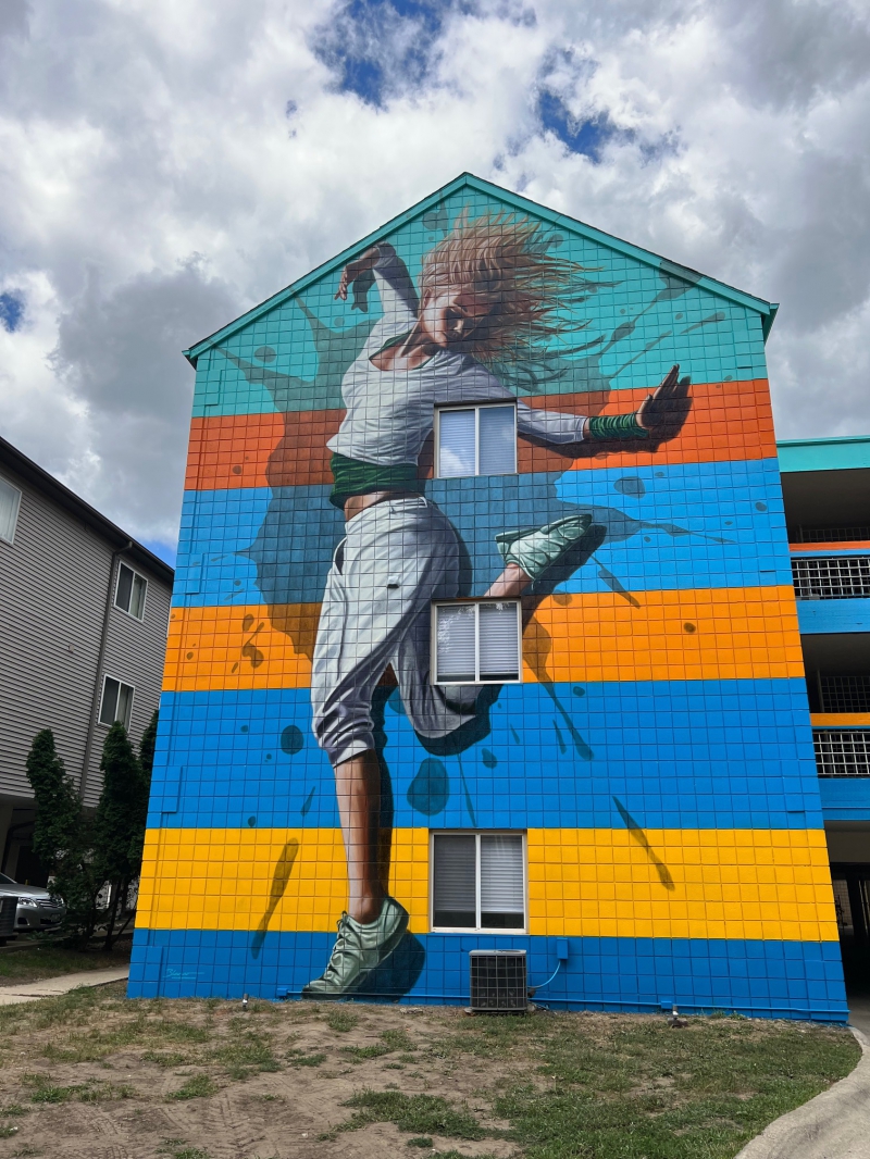 A brightly colored mural covers the entirety of the side of an apartment building. It features a woman in baggy pants and shirt and tennis shoes dancing. Photo by Julie McClure.