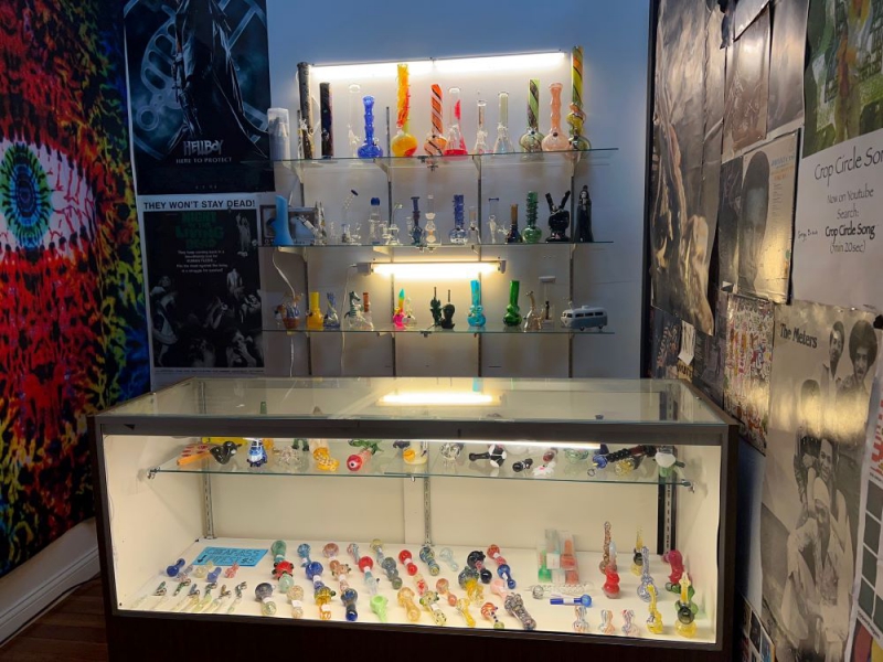 A glass case filled with glassware. Behind the case are wall shelves lined with glass bongs. Tapestries hang on the walls on either side of the room. Photo by Julie McClure.