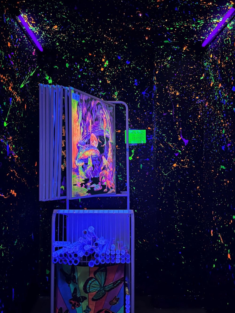 The interior of a room is painted black with spatters of fluorescent paint. There is a rack of posters, and black lights hanging on the ceiling, lighting the room. Photo by Julie McClure.