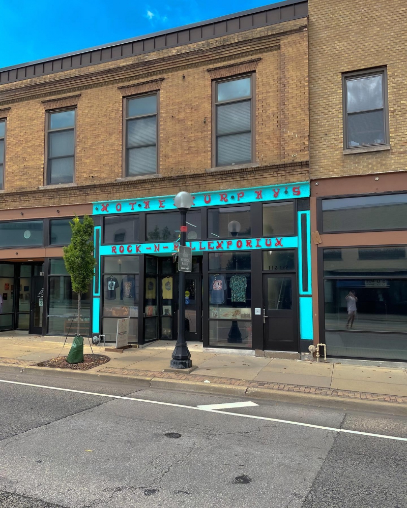 In a row of brown brick facades, there is a turquoise painted store front with red letters that say Mother Murphy's Rock - N - Roll Emporium. There are posters and shirts hanging in the windows. Photo from Mother Murphy's Facebook page.