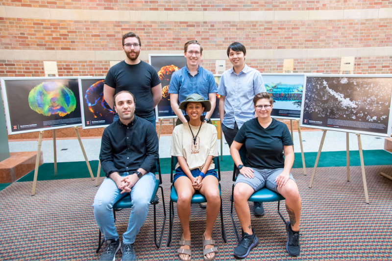Three people are seated in chairs, and three others are standing behind them. They are placed in front of several images on easels. Photo from Beckman Institute.