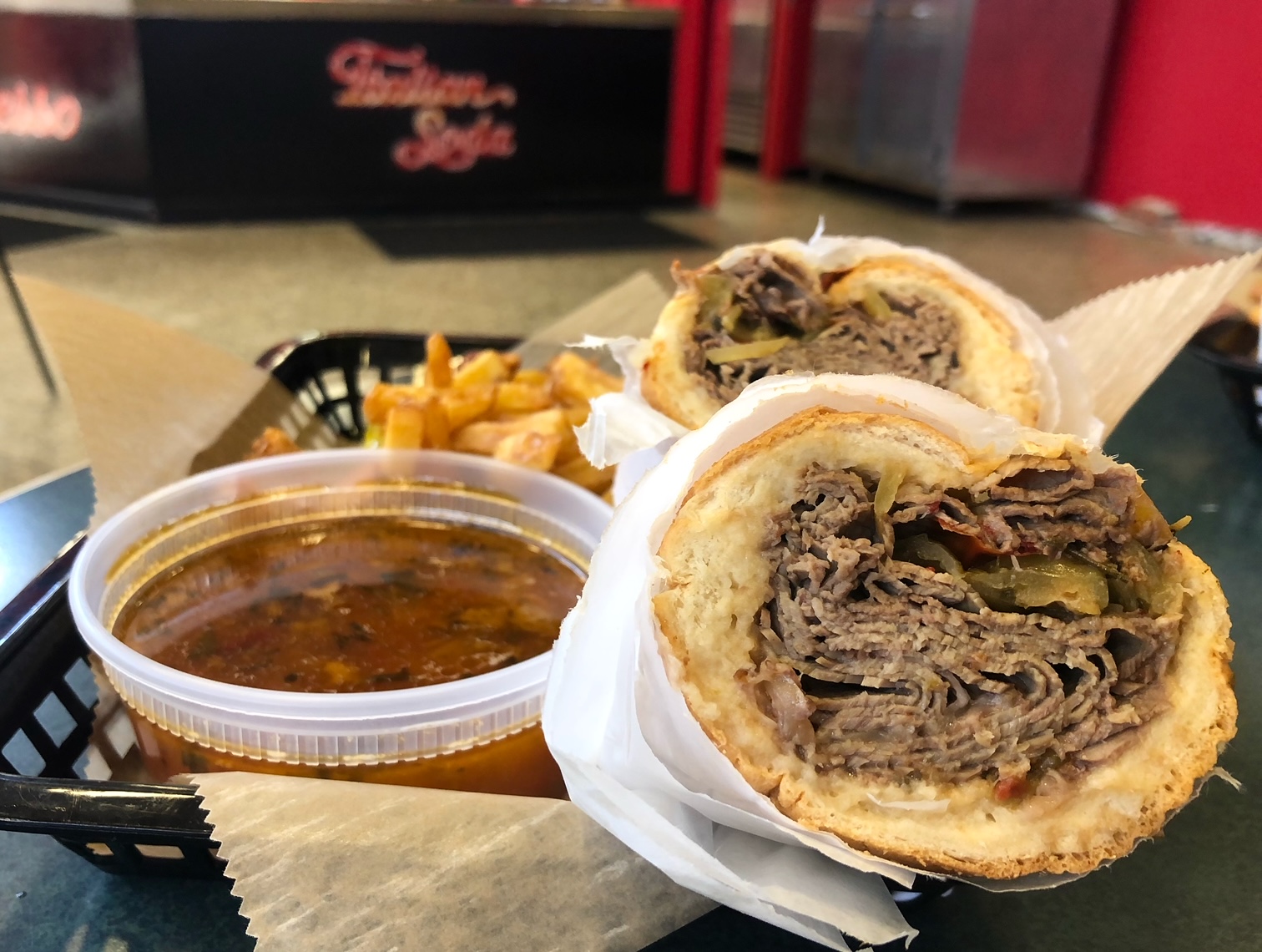 An Italian beef sandwich from Baldarotta's in Urbana, Illinois is sliced in half on a plastic tray. There is a large container of au jus beside it and a side of fries. Photo by Alyssa Buckley.