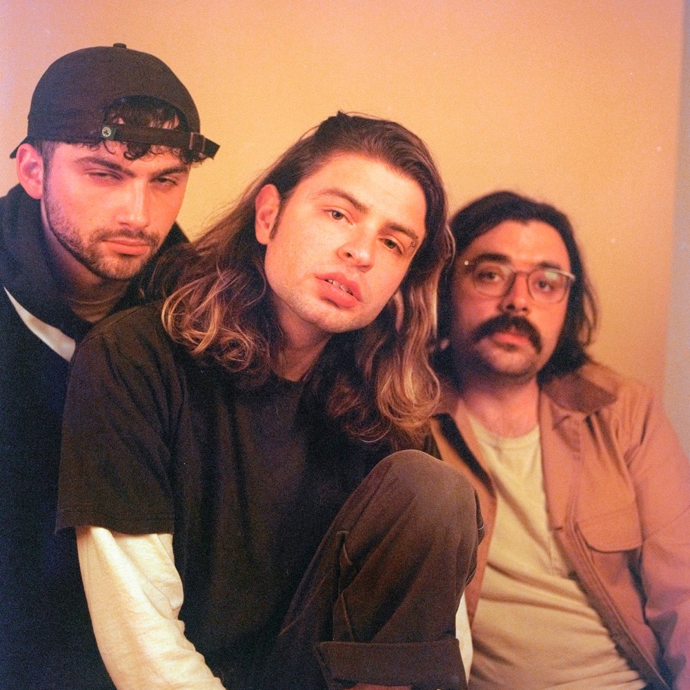 Studio portrait of band Horse Jumper of Love. The three members sit in front of an orange backdrop, looking into the camera.