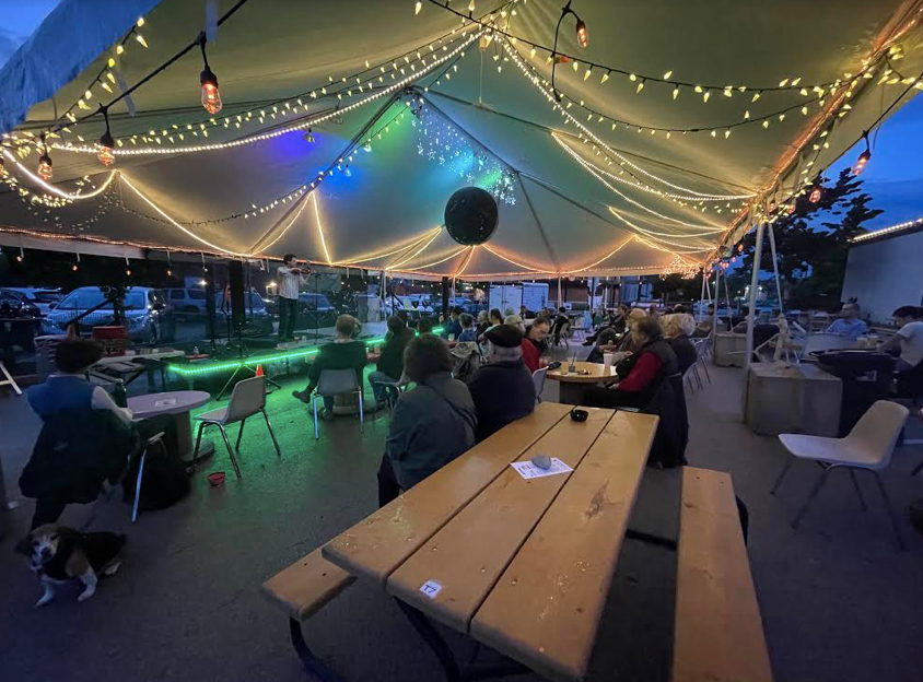Image of the outdoor patio at Rose Bowl Tavern, with string lights surrounding the tent, while onlookers sit at picnic tables and watch a performer. 