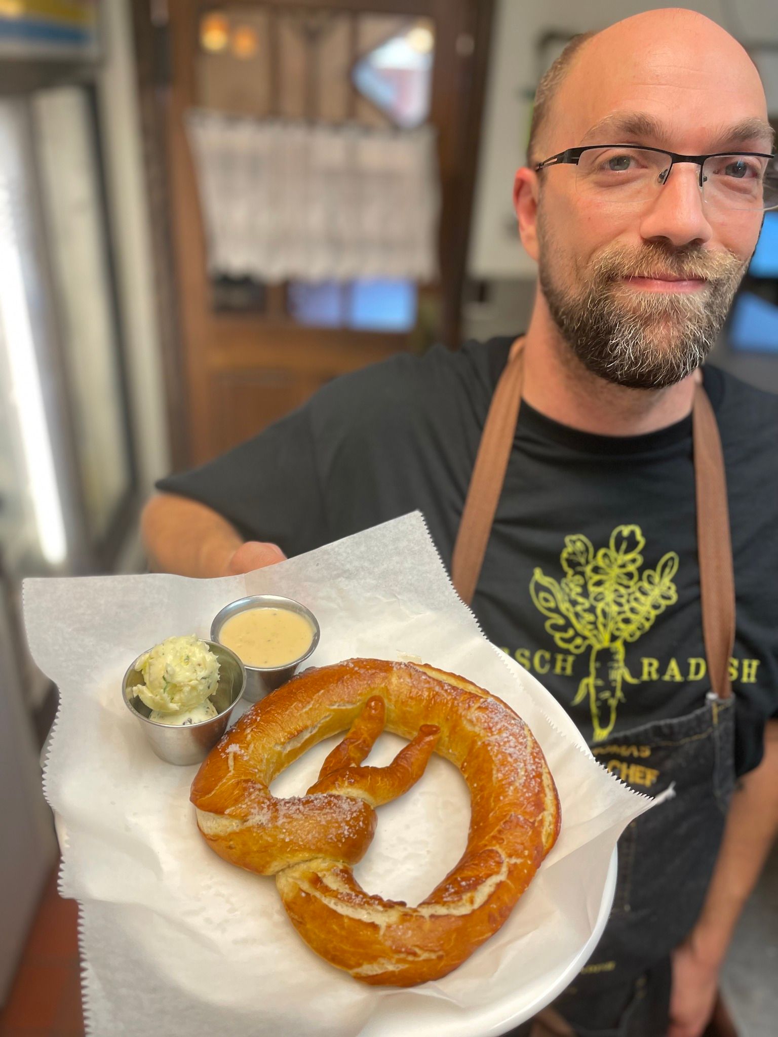 A chef at Horsh Radish in Gibson City, Illinois holds a single pretzel with two cups of dips. Photo by Horsh Radishâ€™s Facebook page.