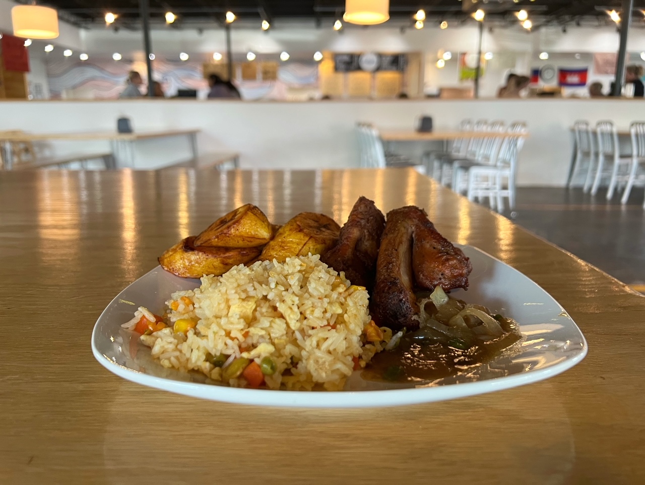 On a wooden table inside Broadway Food Hall in Urbana, there is a white plate of Congolese food: pork ribs, fried rice, and plantains by the restaurant Les Gourmets Cuisine. Photo by Alyssa Buckley.