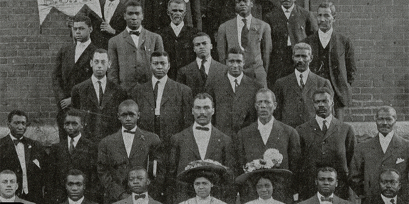 Early 20th Century black and white photo of a community of Black men in suits and Black women in dresses and hats taken against a brick building. 