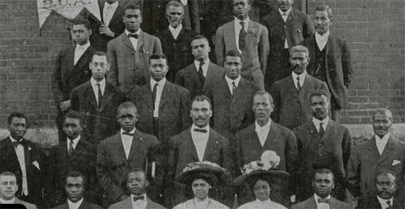 Early 20th Century black and white photo of a community of Black men in suits and Black women in dresses and hats taken against a brick building. 