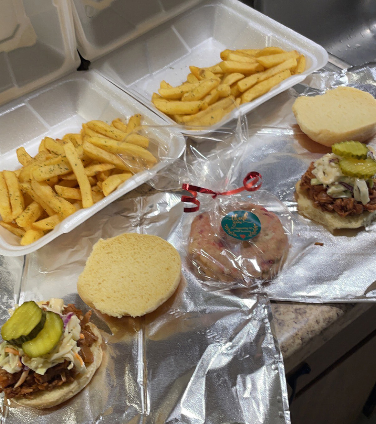 A closely cropped image of two barbcue jackfruit sandwich meals and a strawberry cream packaged cookie in the middle. The photo is slightly out of focus. Pickles, slaw, and BBQ Jackfruit piled on hamburger-style buns are shown open-faced on aluminum foil sheets. In the background are french fries in two styrofoam take-out containers. Photo by Lamiea Wilson.