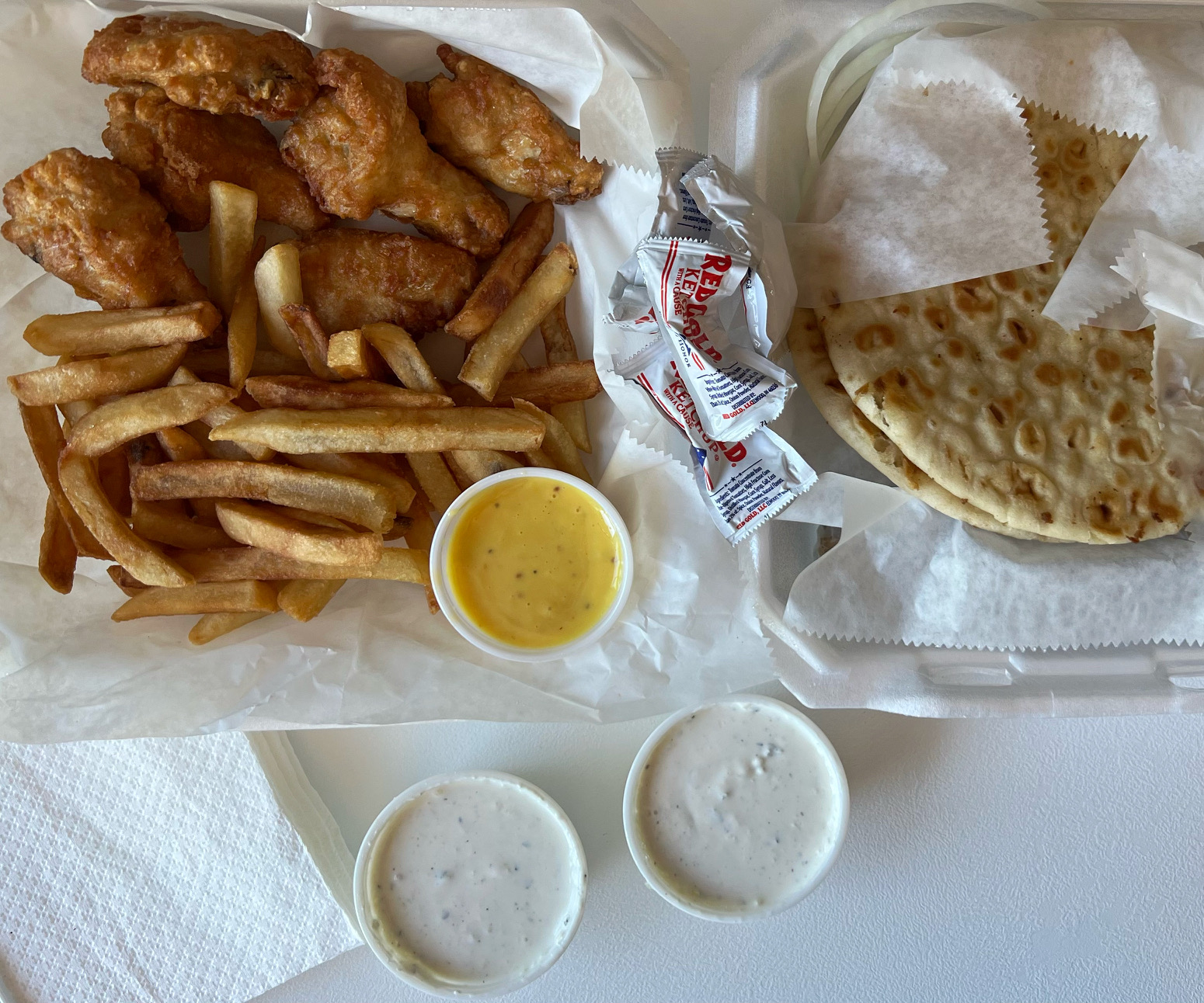 An overhead photo shows the author's lunch. On the left, a parchment paper lined styrofoam container has fried chicken wings with fries and a side sauce of honey mustard. The right has pitas over wrapped gyro meat with two cups of gyro sauce. Photo by Alyssa Buckley.