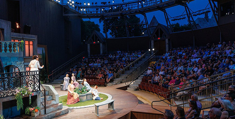 Photo of outdoor theatre in the round under the stars during Illinois Shakespeare Festival. One actor on a balcony and a group of five on the ground below.