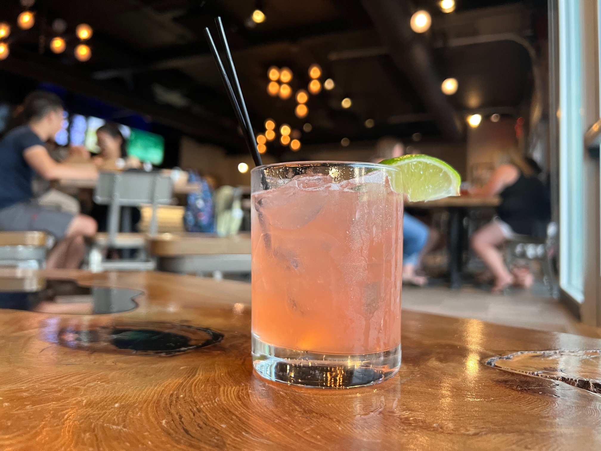 On a wooden table at Collective Pour, a strawberry margarita in a half tumbler is in focus while the other patrons and the lights of the bar are blurred. Photo by Alyssa Buckley.