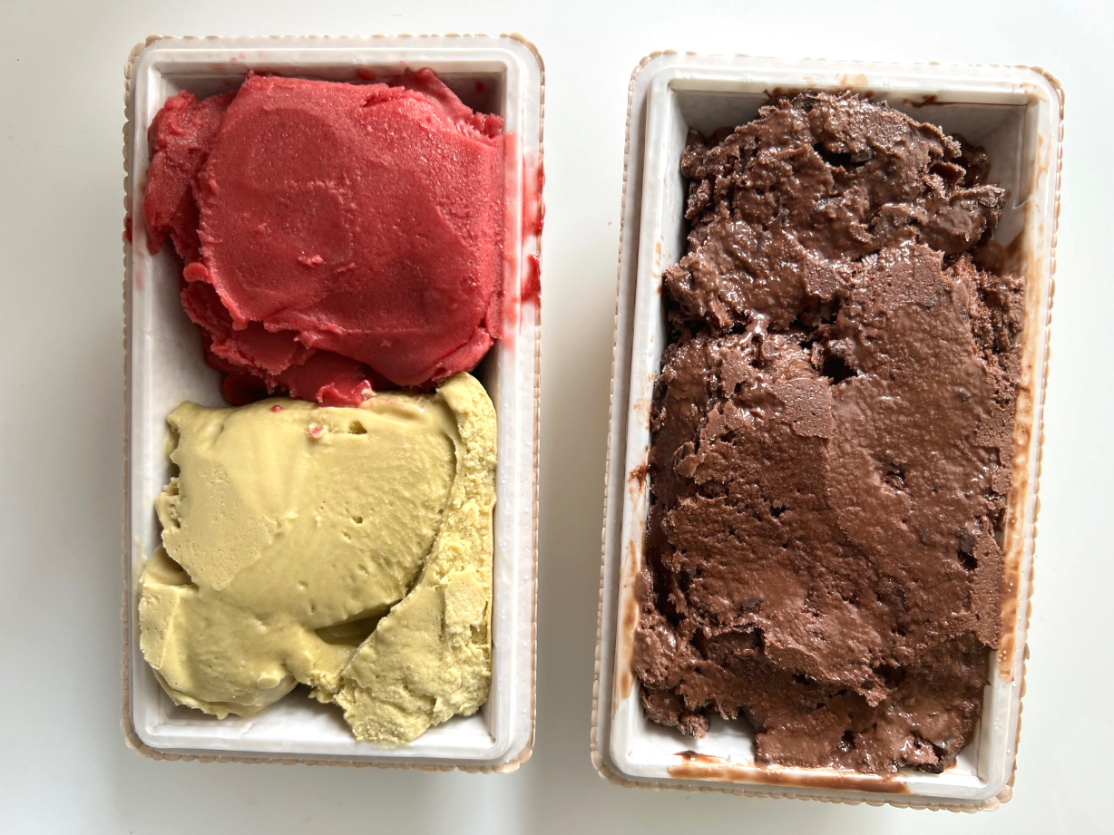 On a white table, there are two rectangular pints of gelato from Baldarotta's. On the left, there is a red raspberry scoop on top and a pale green pistachio scoop on the bottom. On the right, the pint is full of dark chocolate gelato. Photo by Alyssa Buckley.
