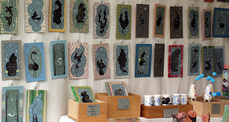 A collection of small fantasy creatures painted in black ink against blue cloud like backgrounds mounted on small patterned rectangles hung from the tent wall.
