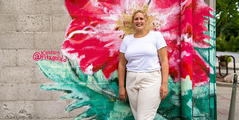 Muralist Kinsey Fitzgerald posing against  the Rose Bowl Tavern wall in front of a prairie rose with her signature to the left.