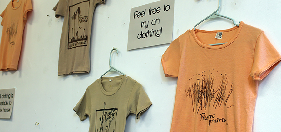 A selection of t-shirts saying preserve the prairie in earth tones hanging on a white wall. 
