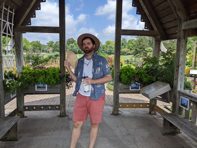 A man in a beige hat, white t-shirt, blue collared shirt, and red shorts is standing in the center of a gazebo, holding a water bottle and fanning himself with a small fan. Photo by Andrea Black.