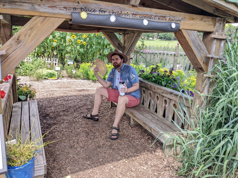 A man in a beige hat, white t-shirt, blue collared shirt, and red shorts is sitting on a wooden bench and there are wooden beams overhead. He is surrounded by a garden of flowers. Photo by Andrea Black. 