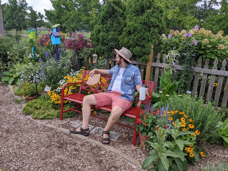A man in a beige hat, white t-shirt, blue collared shirt, and red shorts is sitting on a red metal bench holding a fan and water bottle. He is surrounded by a flower garden with colorful bird houses. Photo by Andrea Black.