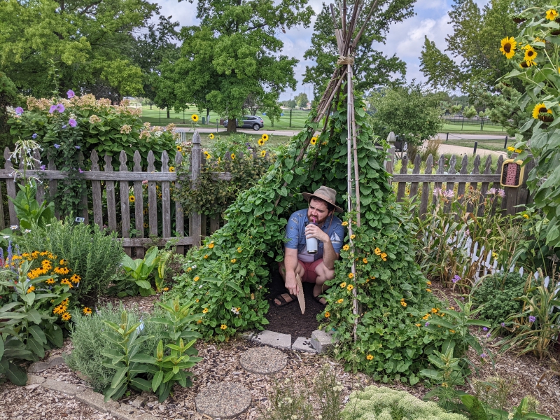 A man in a beige hat, white t-shirt, blue collared shirt, and red shorts is crouched under a small canopy of flowers and vines. Photo by Andrea Black. 