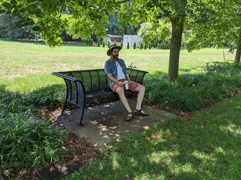 A man in a beige hat, white t-shirt, blue collared shirt, and red shorts is sitting in the shade on a black metal bench on a concrete slab. Photo by Andrea Black.