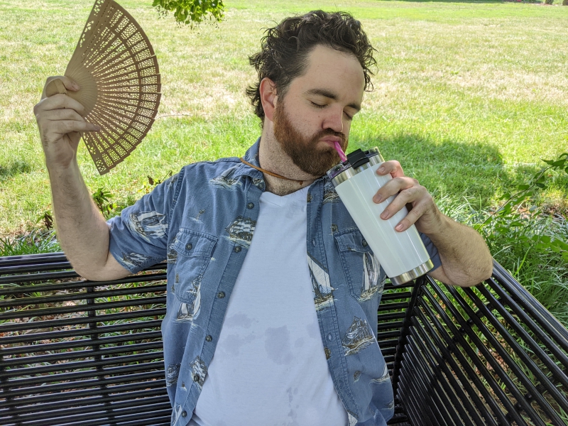 Close up of a man in a white t-shirt and blue collared shirt sipping a water bottle and fanning himself. Photo by Andrea Black.