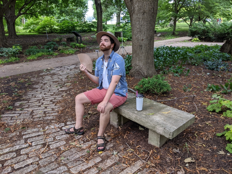 A man in a beige hat, white t-shirt, blue collared shirt, and red shorts is sitting on a stone bench in the middle of a grove of trees, fanning himself. Photo by Andrea Black.