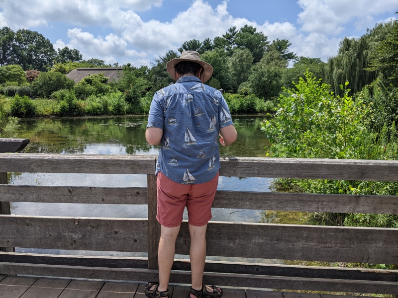 A man in a beige hat, white t-shirt, blue collared shirt, and red shorts is standing on a wooden bridge, facing away from the camera, looking into a pond. Leafy green trees surround the pond. Photo by Andrea Black.