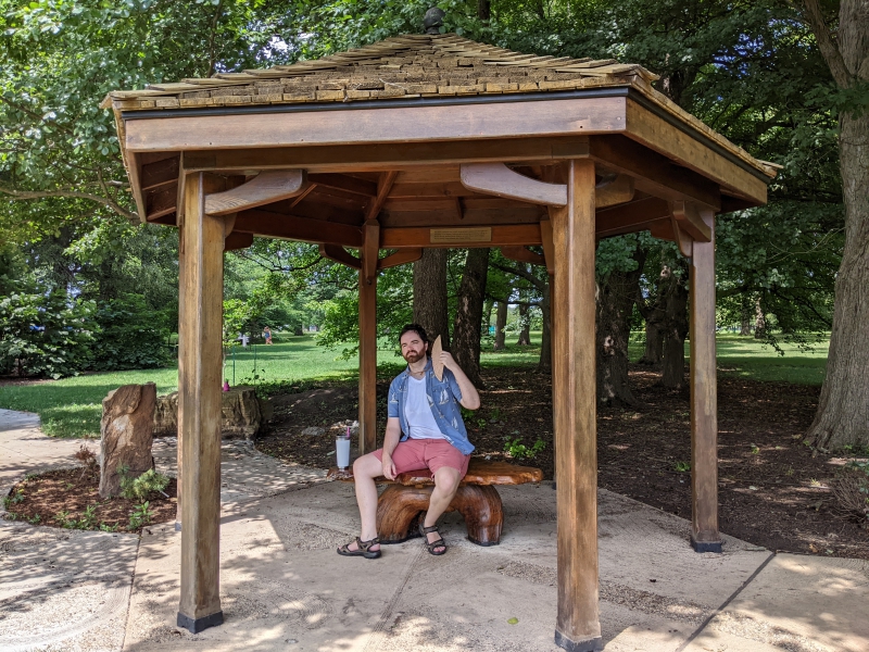 A man in a white t-shirt, blue collared shirt, and red shorts is siting on a sculpted wooden bench under a wooden gazebo fanning himself. Photo by Andrea Black.