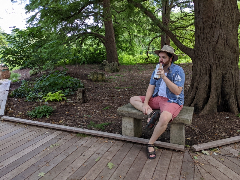 A man in a beige hat, white t-shirt, blue collared shirt, and red shorts sits on a stone bench sipping from a bottle of water. A planked pathway runs in front of him. Photo by Andrea Black. 